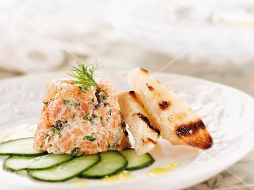 Smoked Trout Tartare with Sliced Cucumbers and Grilled Italian Bread Slices on a White Plate