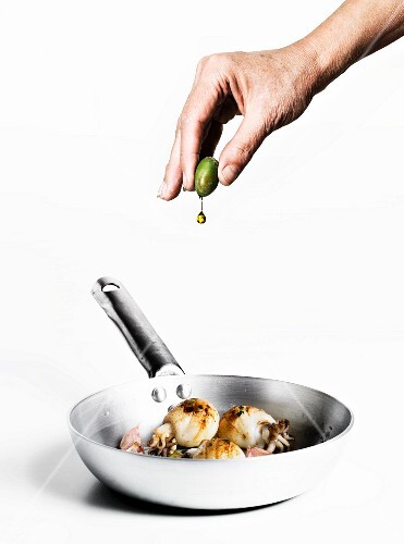A hand squeezing olive oil from an olive onto sepia with garlic