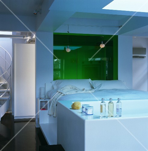The white platform bed in this open-plan bedroom has a bath positioned at the end of it and wardrobes behind the green glass panel
