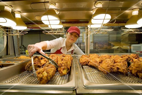 Woman Selecting Piece of Fried Chicken From Under Heat Lamps