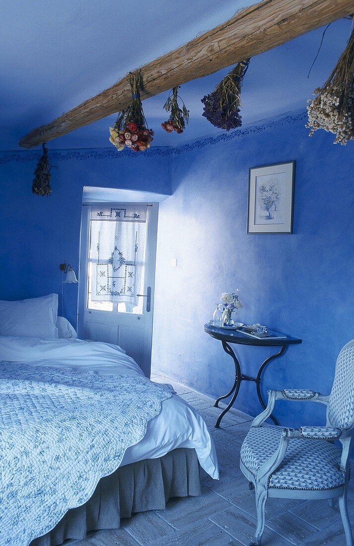 Blue bedroom with dried flowers hanging from ceiling beams; floral, quilted counterpane on double bed