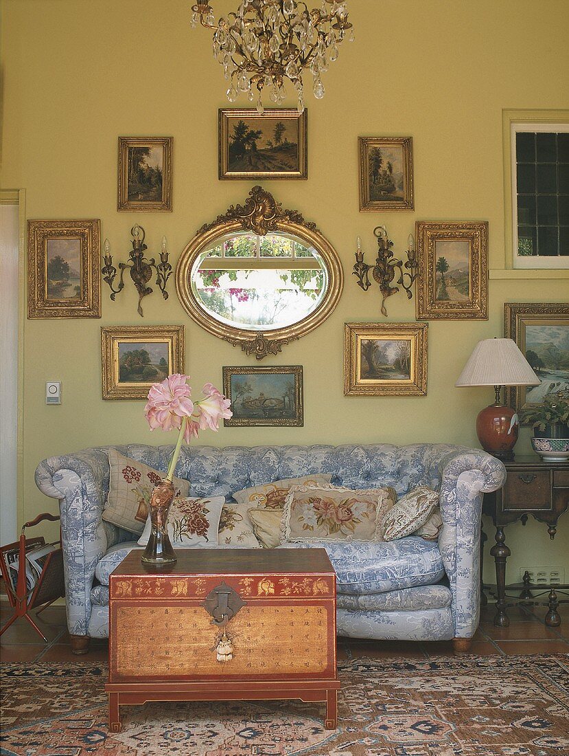 Geblümtes Sofa und antike Holztruhe vor antiken Gemälden und einem barocken Spiegel an der Wand