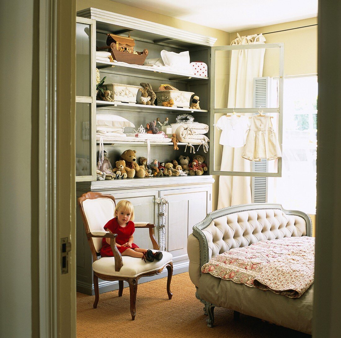 Kleines Mädchen sitzt auf Stuhl vor Schrank in Kinderzimmer