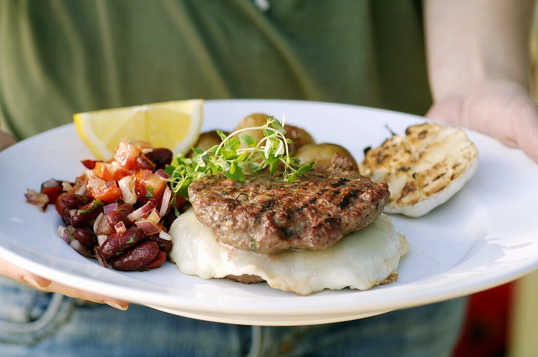 Person hält Grillteller mit Burger und Tomaten-Bohnen-Salat