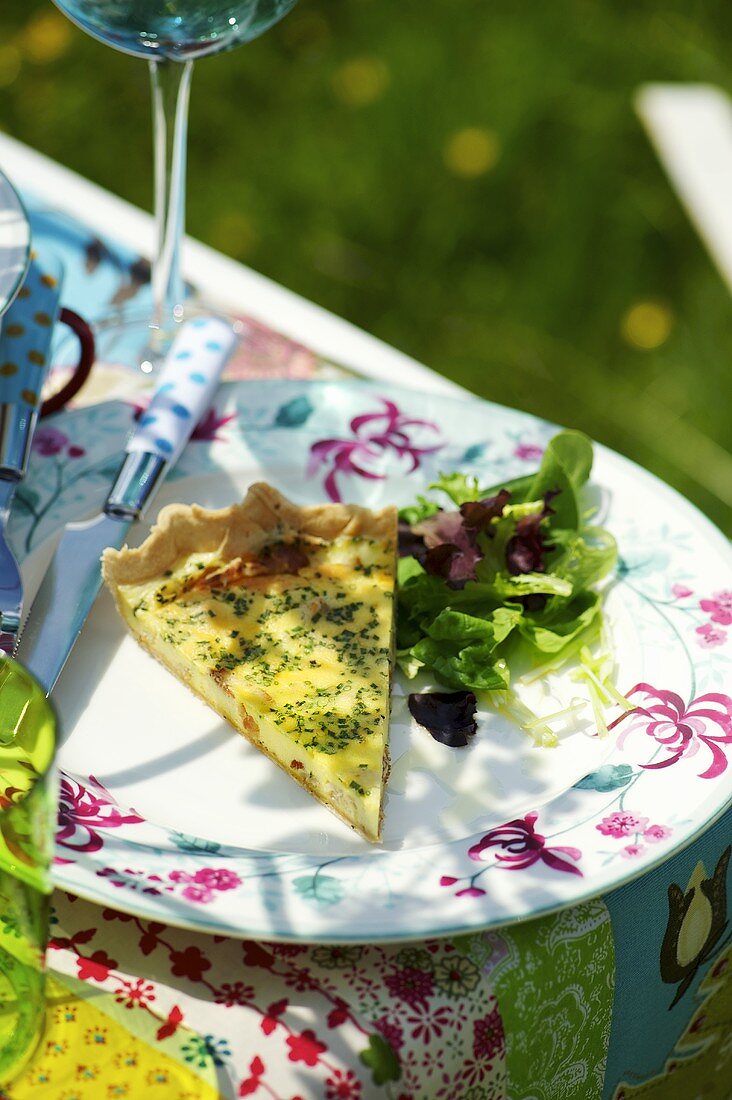 Ein Stück Käsequiche mit Blattsalat auf Gartentisch