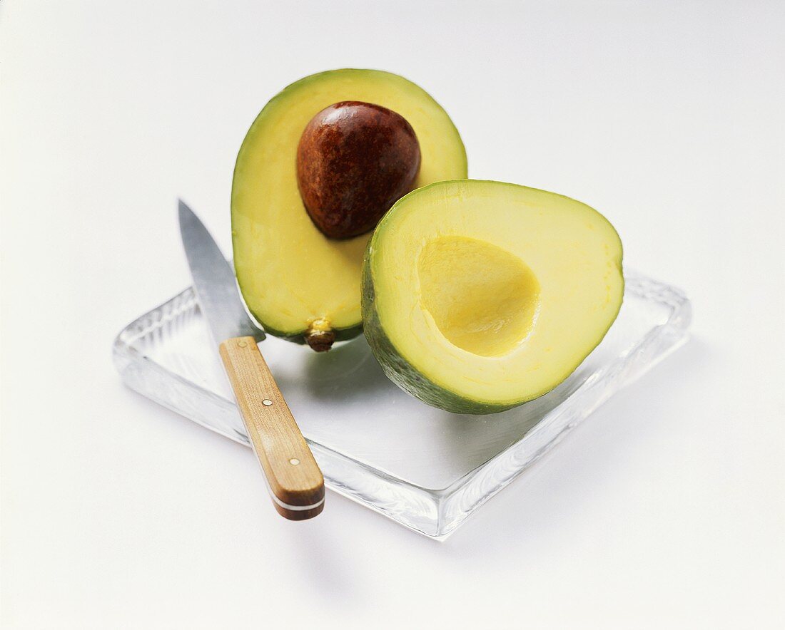 A halved avocado with knife on a glass plate