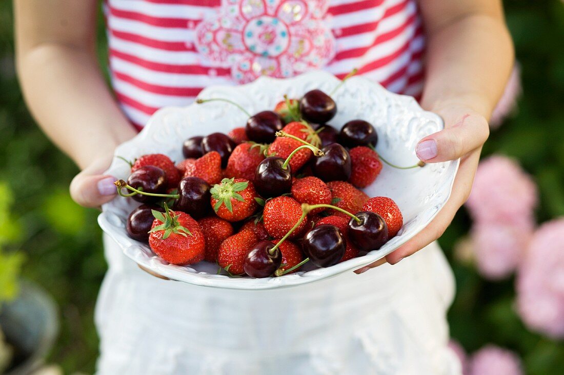 A child holding a white bowl of berries and cherries