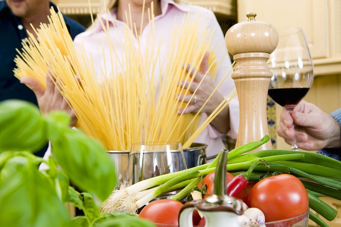 Woman putting spaghetti into pan, herbs & vegetables in front