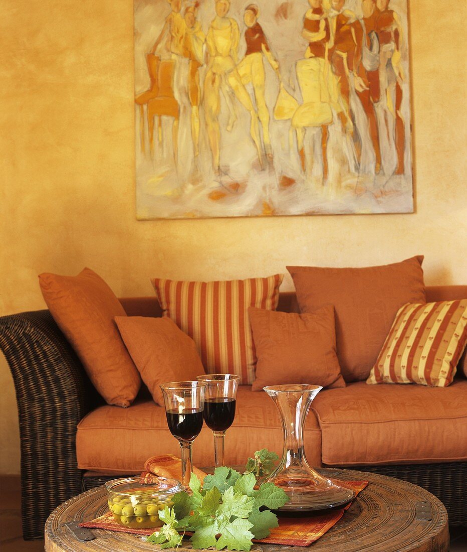 Eingelegte Oliven & Rotwein auf rundem Couchtisch vor Rattansofa mit roten Kissen in Wohnzimmer