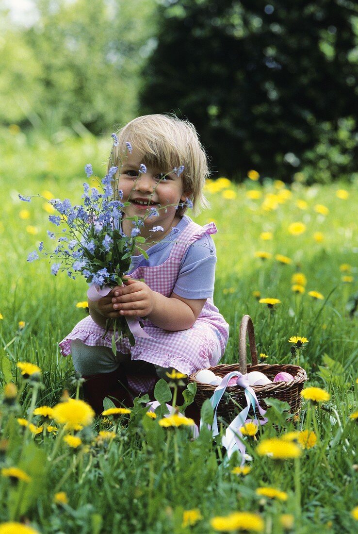 A little girl holding flowers with a basket of Easter eggs next to her