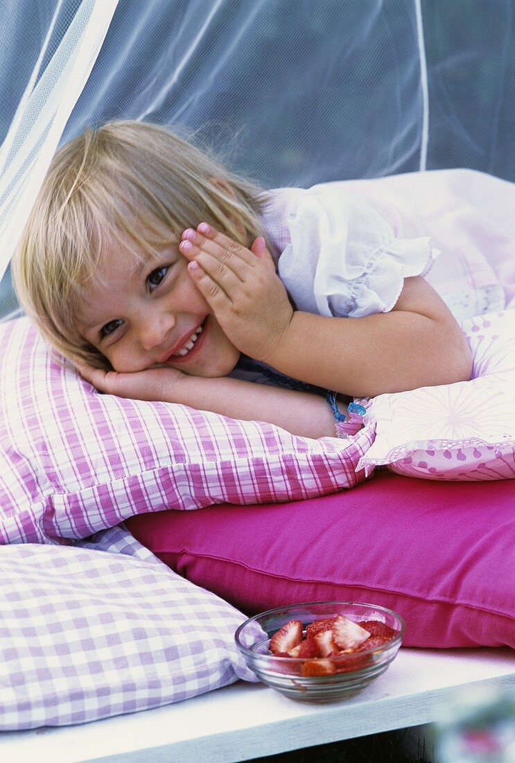 Kleines Mädchen liegt auf Kissen neben einer Schale Erdbeeren