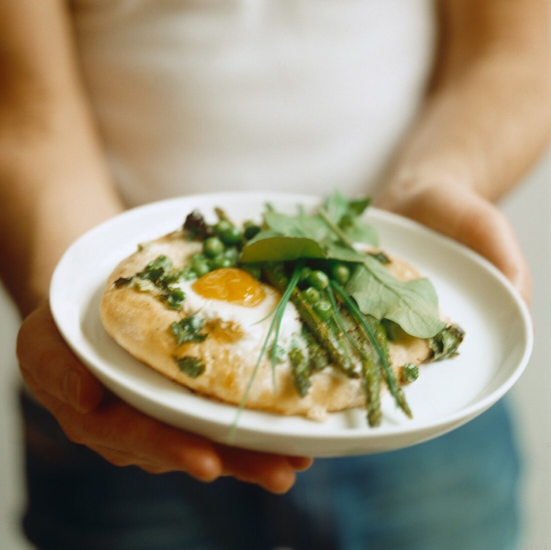 A man holding a pizza topped with egg, peas, asparagus and spinach