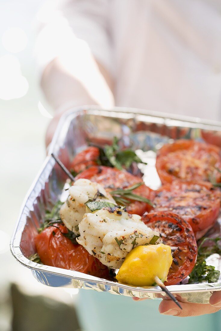 Frau hält Aluschale mit gegrilltem Fischspiess und Tomaten