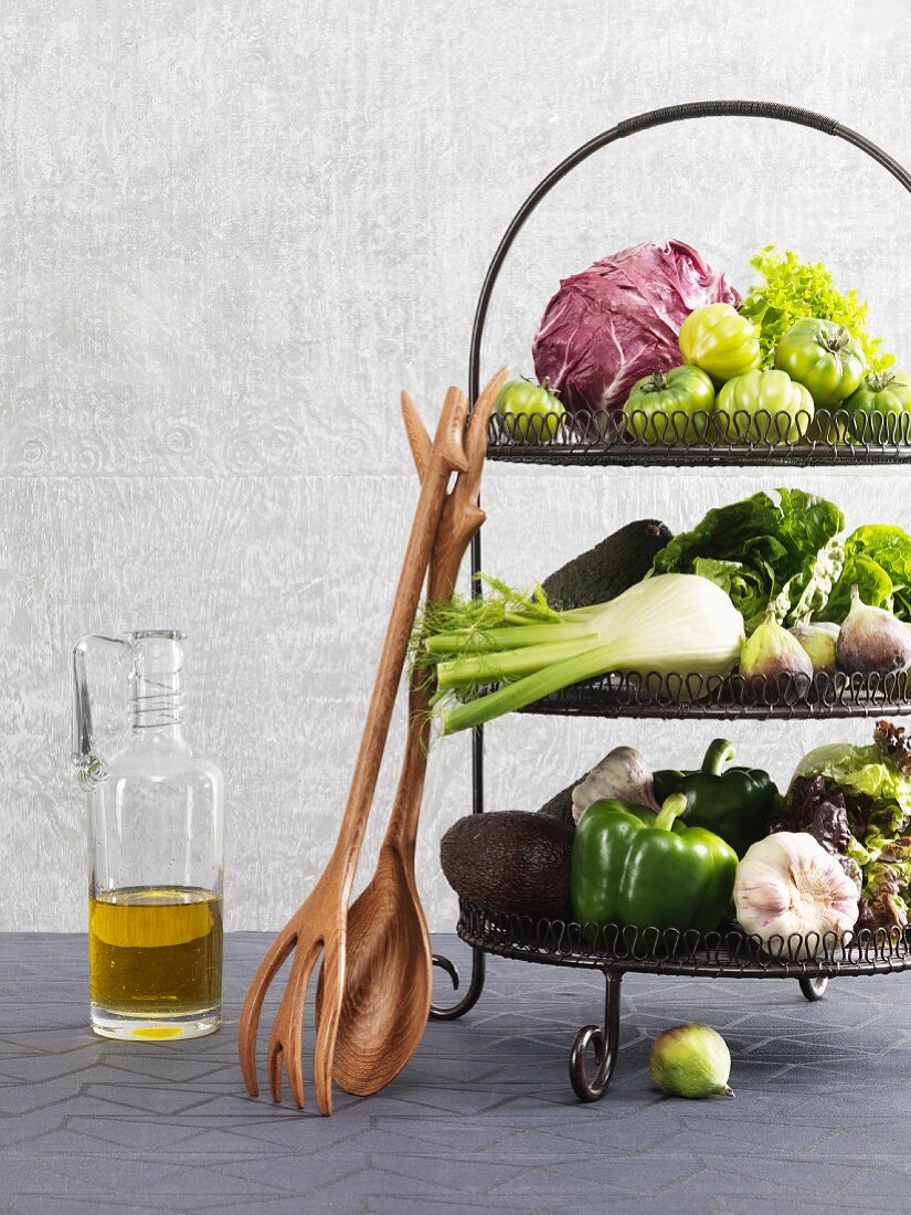 Vegetables on an etagere with wooden salad servers leaning against it and a bottle of oil standing next to it