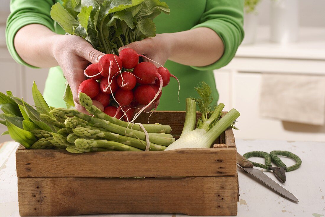 A box of vegetables and a hand holding a bunch of radishes
