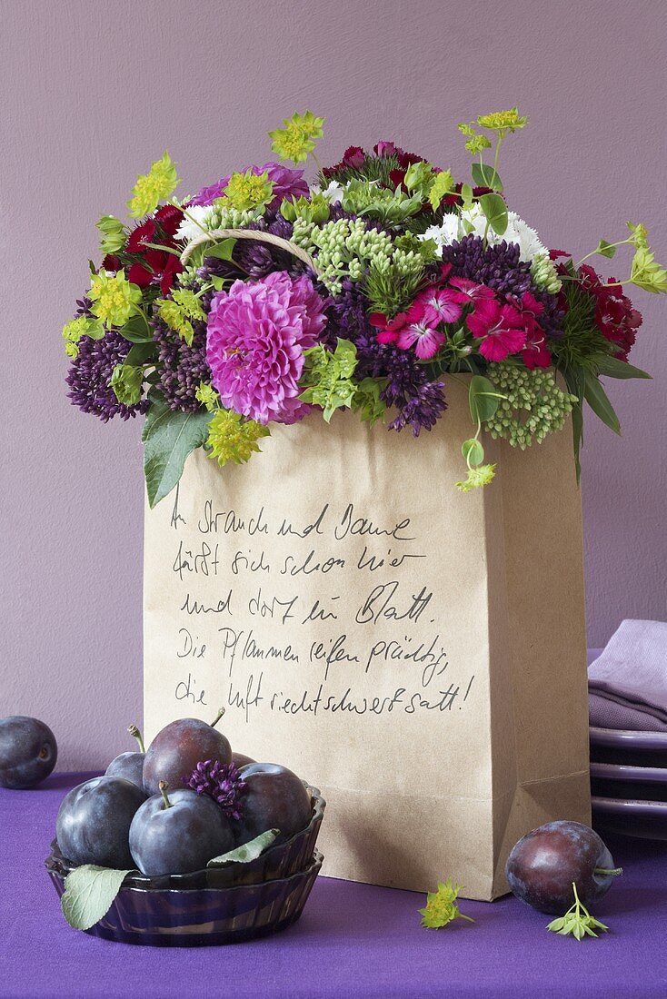 A bouquet of dahlias and stonecrops in a paper bag with plums in front