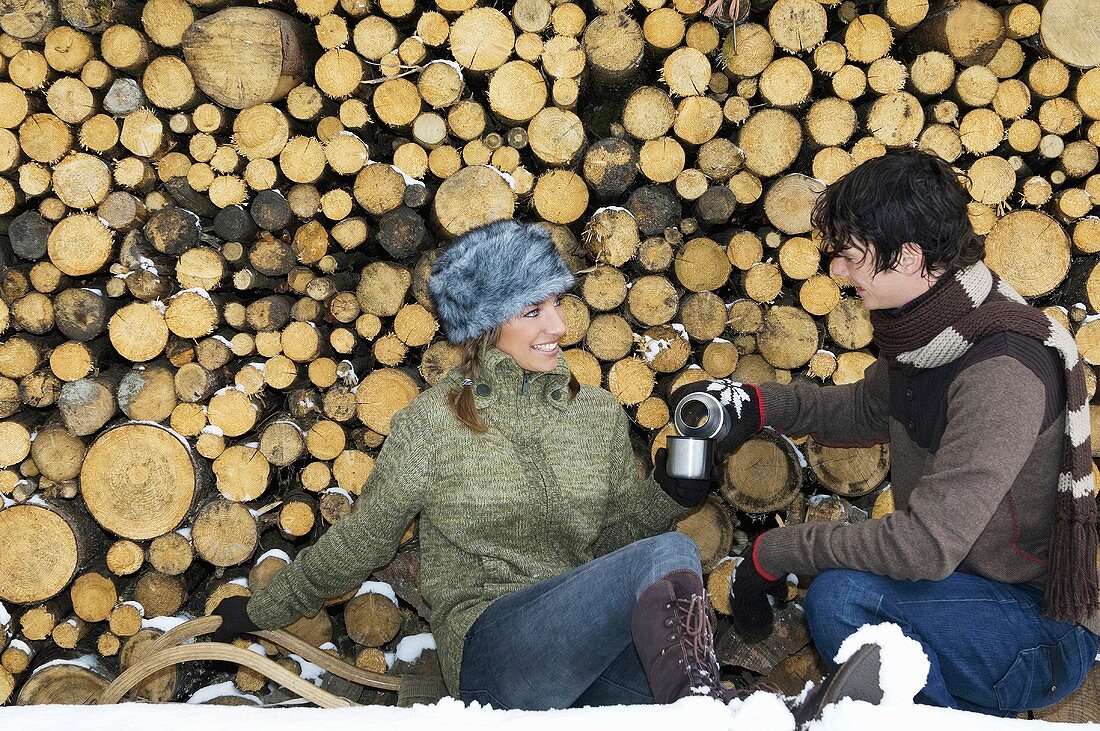 Young couple sitting by stack of wood with Thermos flask