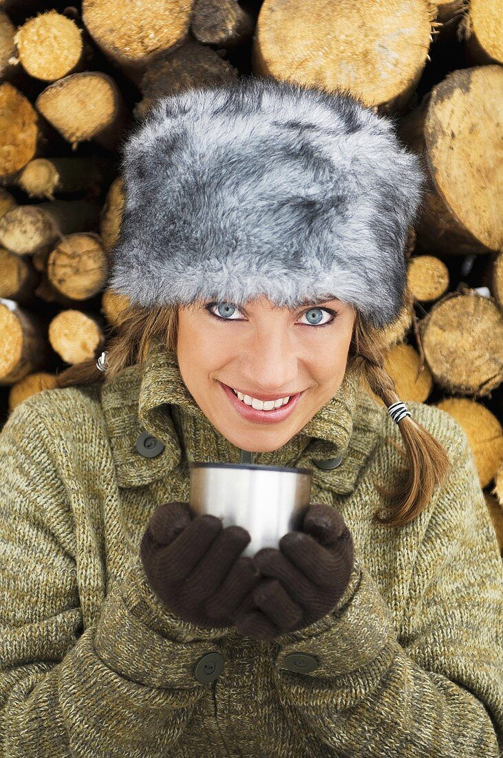 Junge Frau mit Wintermütze und Becher am Holzstapel