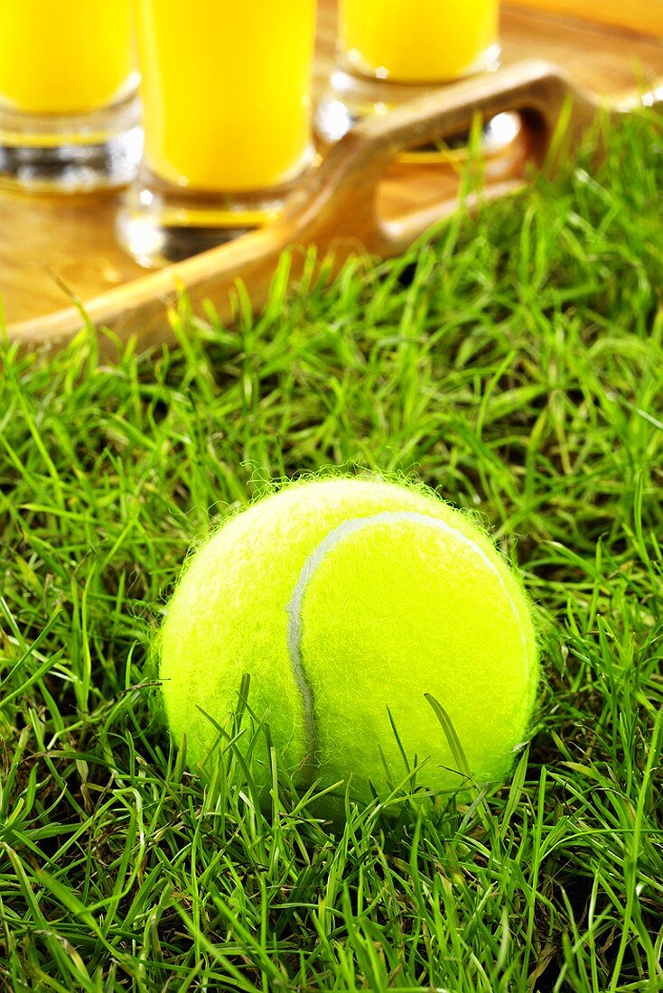 A tennis ball in grass, tray of orange juice behind