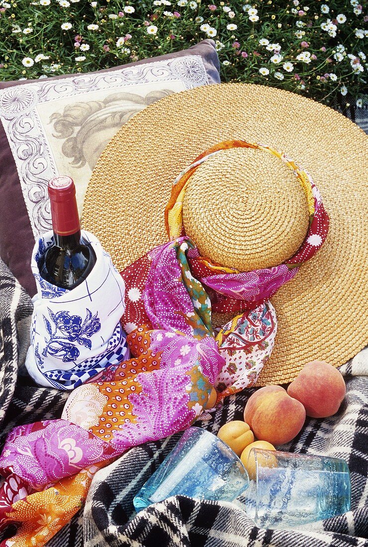 A bottle of red wine, fruit and straw hat on a picnic cloth