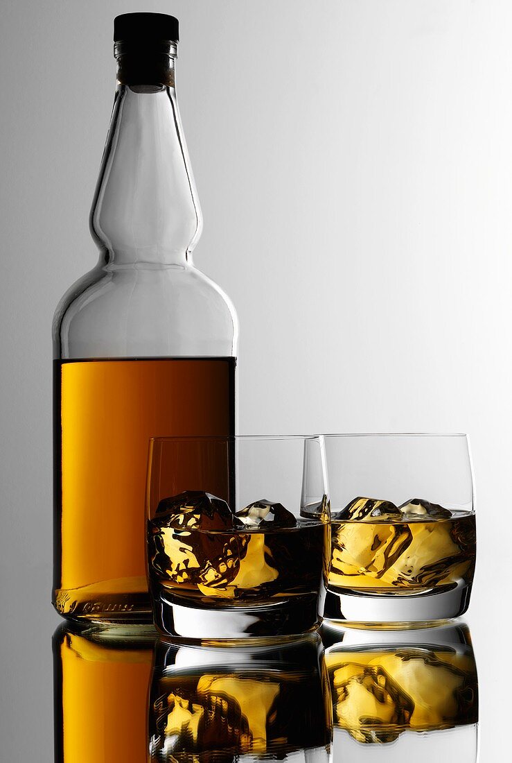 Two glasses of whisky with ice cubes in front of bottle