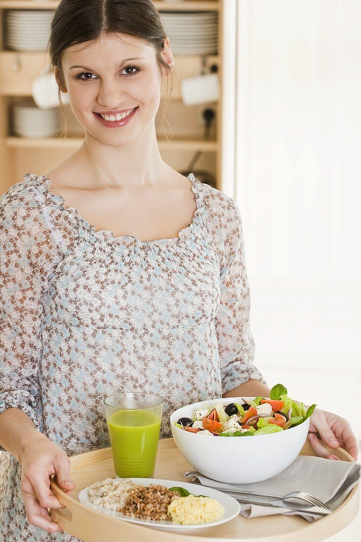 Junge Frau hält Tablett mit Fruchtsaft, Salat und gekochten Getreidesorten