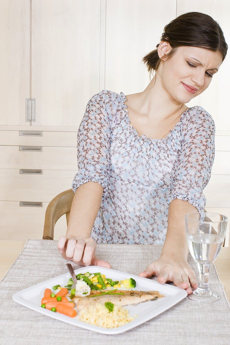 Frau schiebt Teller mit Fisch, Couscous und Gemüse von sich