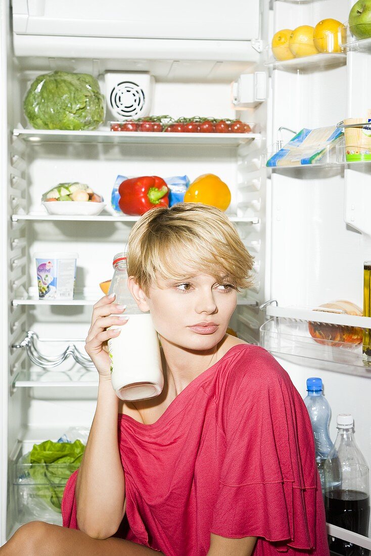 Junge Frau mit Milchflasche sitzt vor geöffnetem Kühlschrank