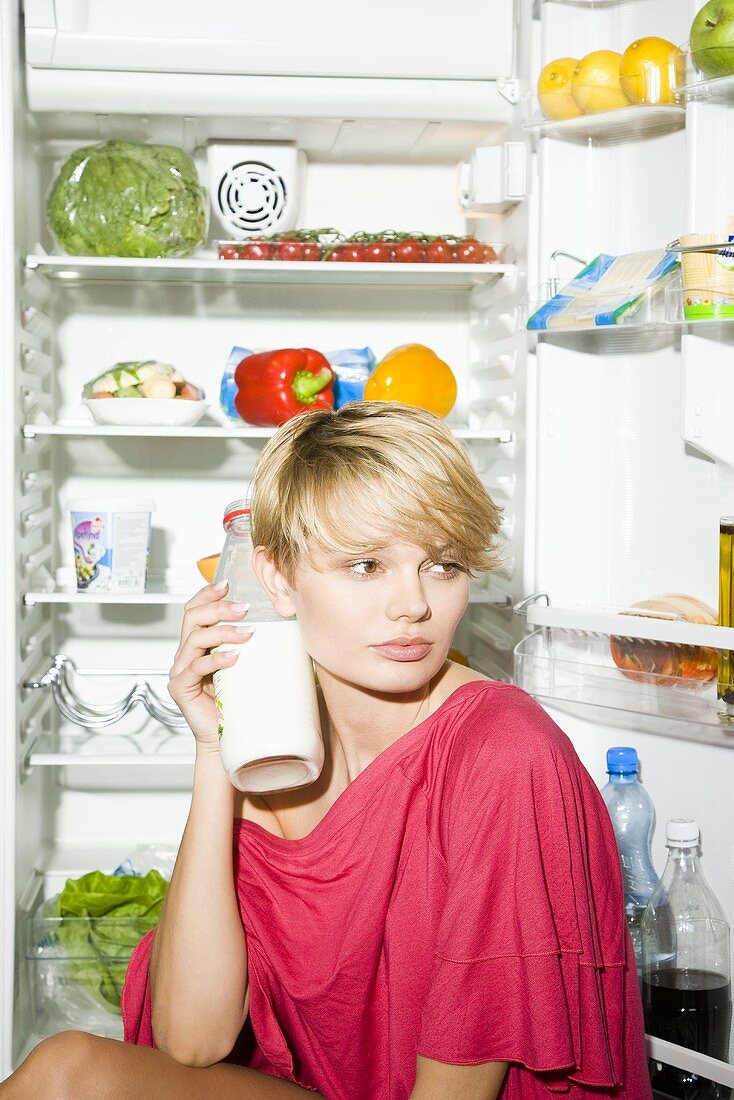 Young woman with bottle of milk sitting in front of opened fridge