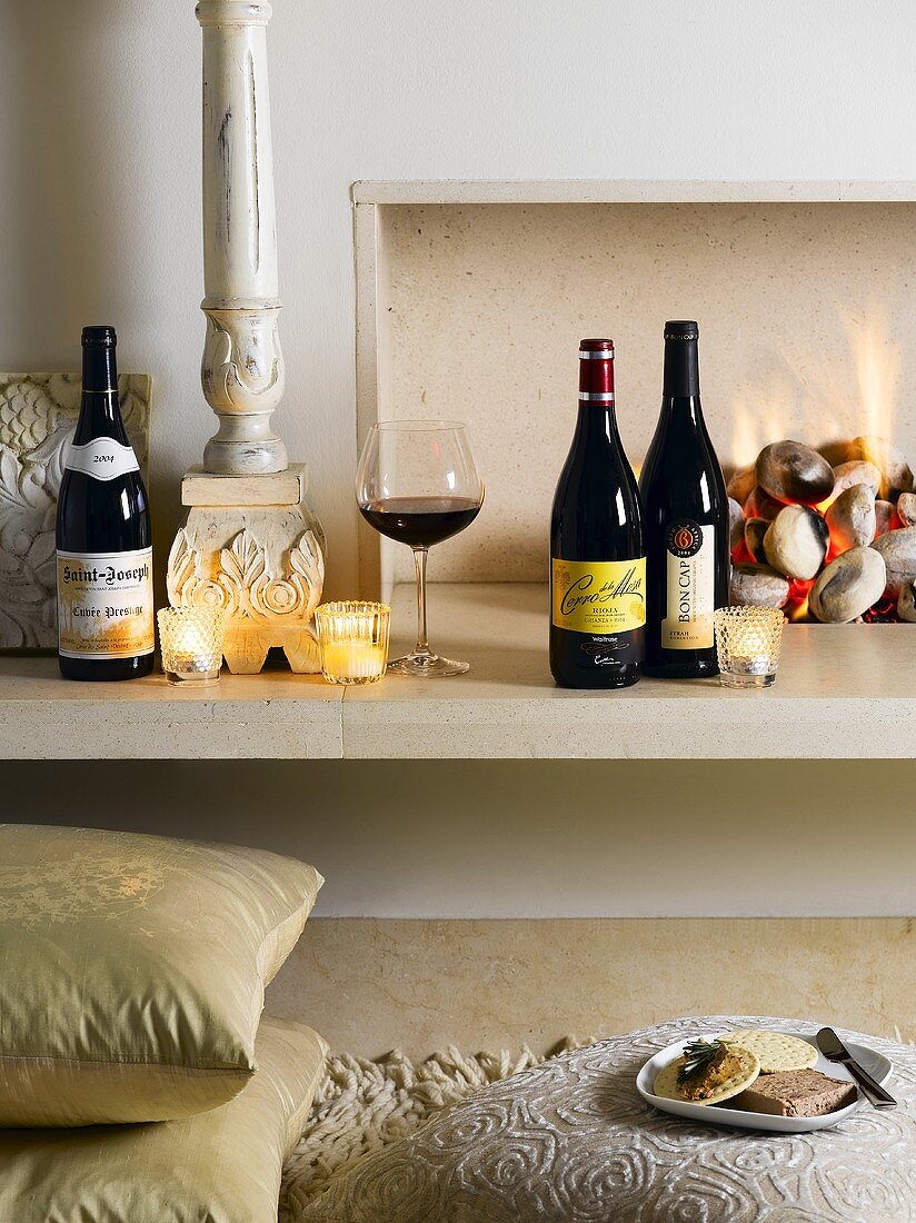 Bottles of red wine, glass of wine and tealights in front of fire