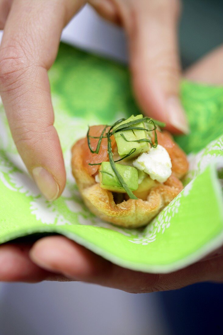 A hand holding a napkin with a salmon and avocado tartlet