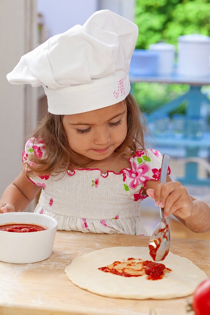 A little girl spreading tomato sauce onto pizza dough