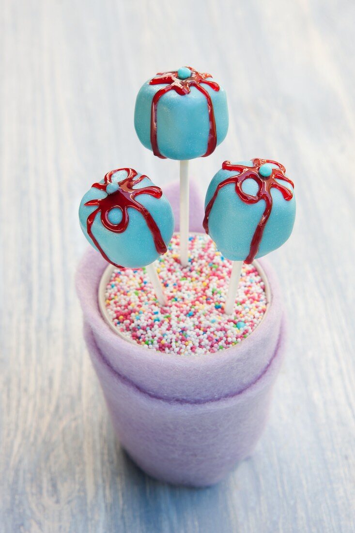 Blue gift-shaped cake pops with red sugar bows in a cup of colourful sugar strands