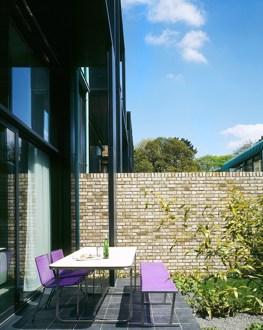 Terrassenplatz mit Stühlen und Bank mit violetter Sitzflächevor Glasfront und Ziegeltrennwand im Garten