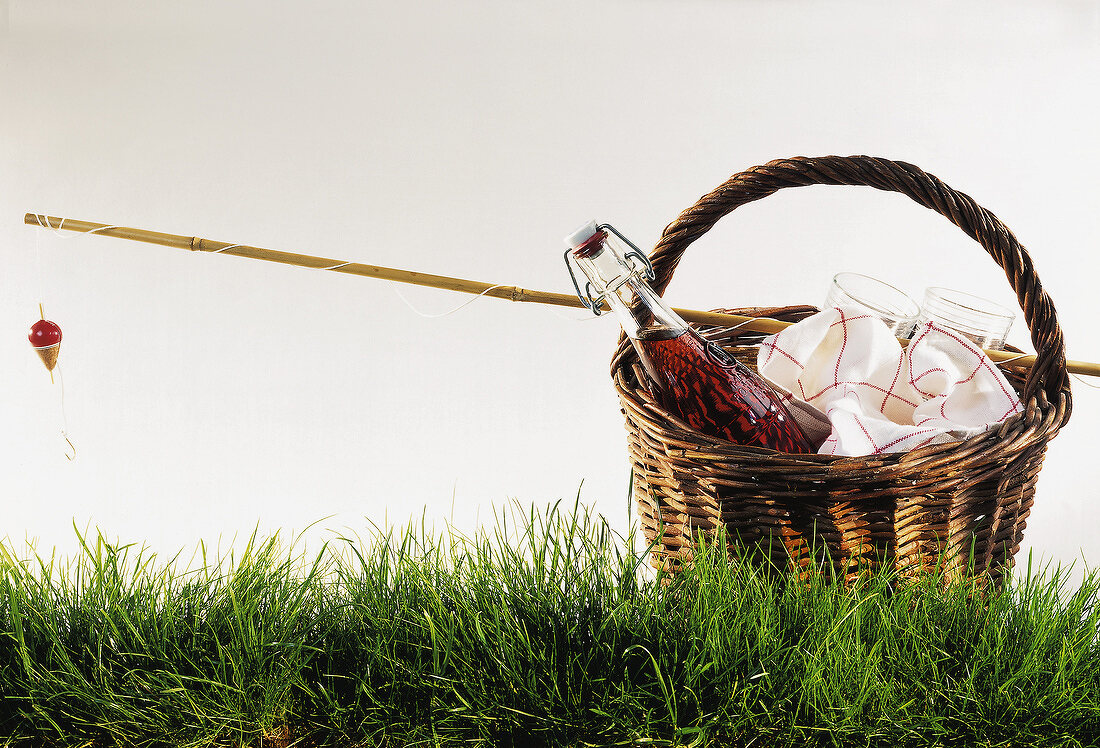 Picknick-Korb mit Rotweinflasche, Gläsern und Angelrute auf Gras