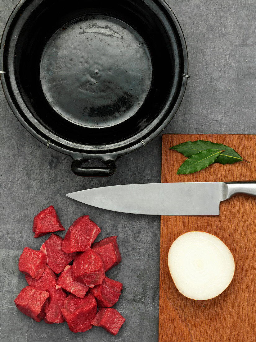 Gewürfeltes Rindfleisch mit Zwiebel, Lorbeerblatt, Messer und Schneidebrett neben einem Topf