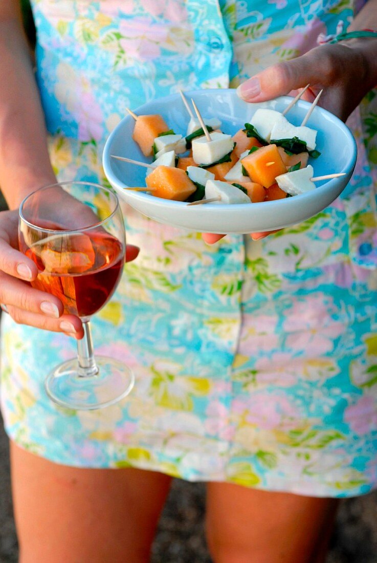 Frau hält einen Teller mit Melonen-Mozzarella-Basilikum-Spiesschen und ein Glas Roséwein