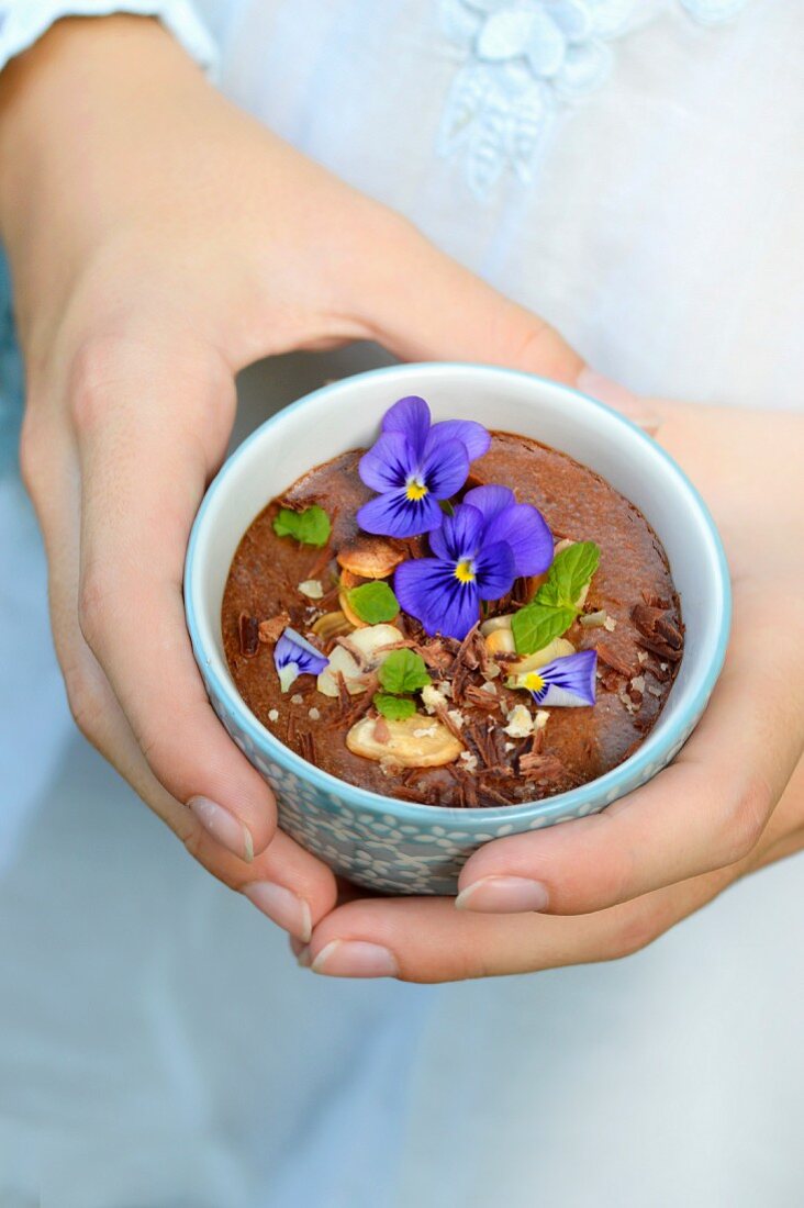 Person holding a chocolate mousse with almonds, hazelnuts, pansies and mint