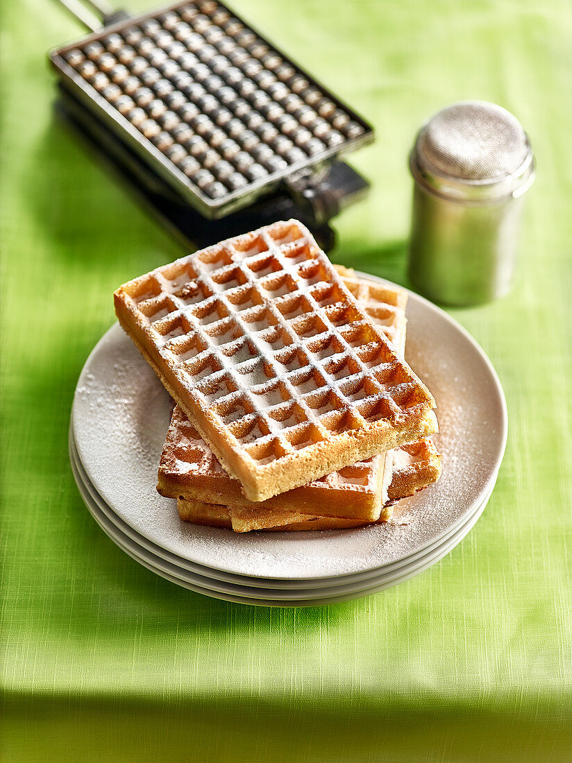 Pile of Brussels waffles sprinkled with icing sugar and a waffle iron