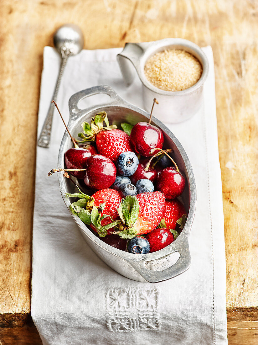 Kirschen, Erdbeeren und Heidelbeeren in silbernem Schmortopf, Tasse mit Rohrzucker