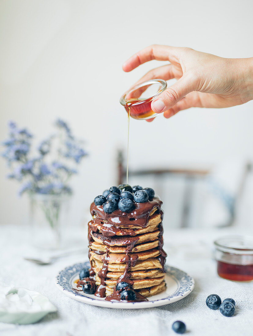 Pouring maple syrup over a pile of pancakes with blueberries and chocolate