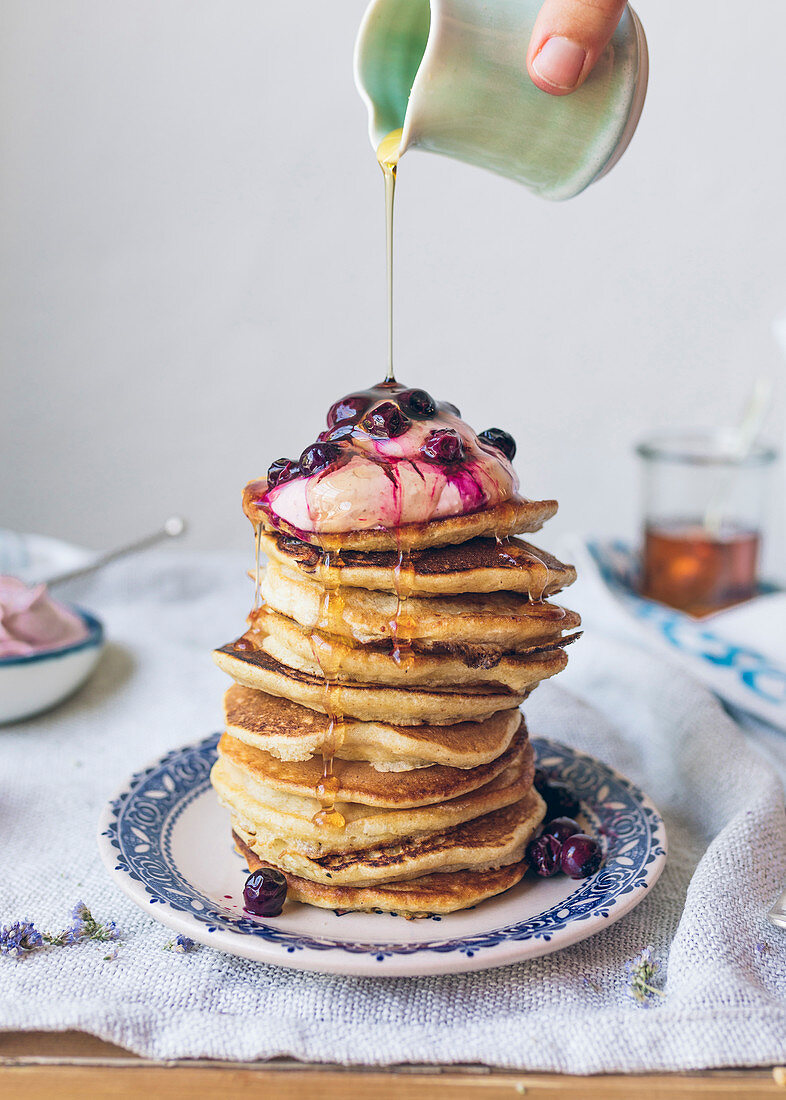Pouring syrup over a pile of pancakes topped with ricotta and blackcurrants