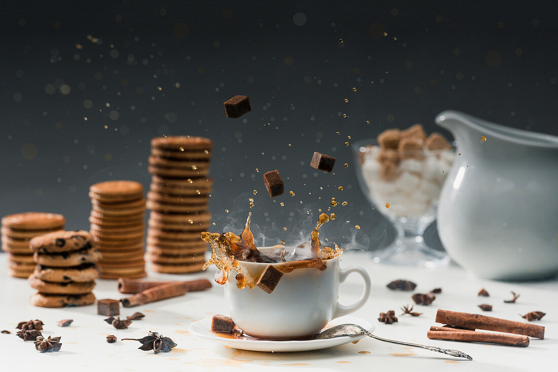 Cane sugar cubes splashing in cup with hot black coffee in front of biscuits and spices