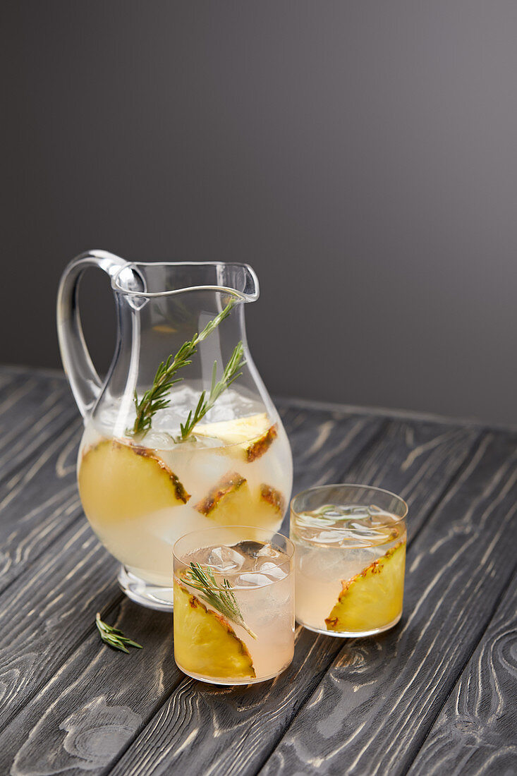jug and two glasses of lemonade with pineapple pieces, ice cubes and rosemary