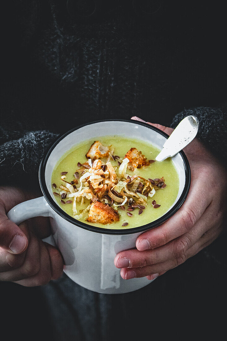 Hands holding enamel cup of leek soup with croutons