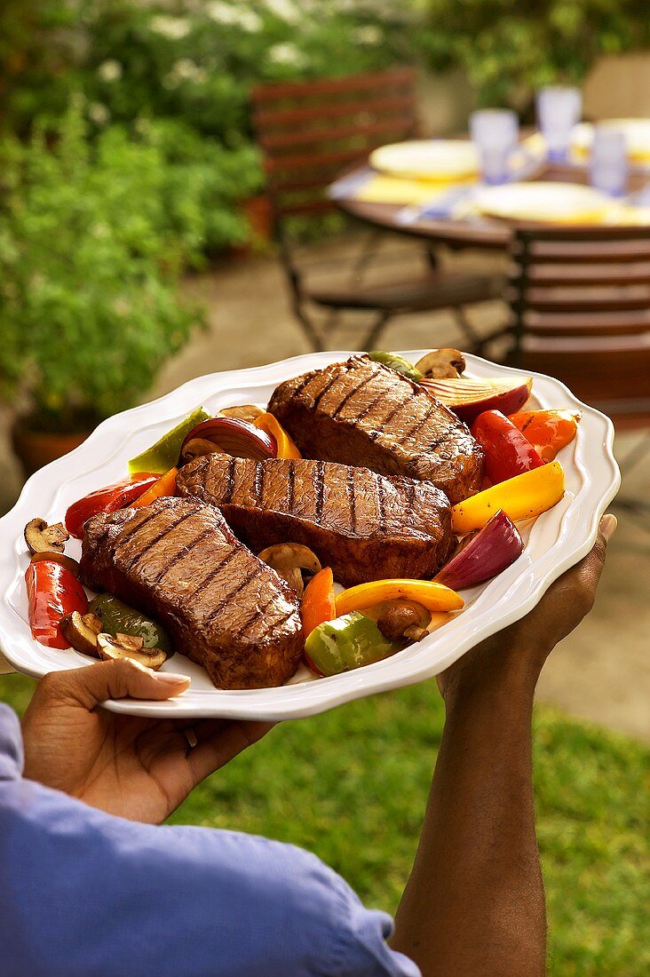 Mann trägt Platte mit gegrillten Steaks zum Gartentisch