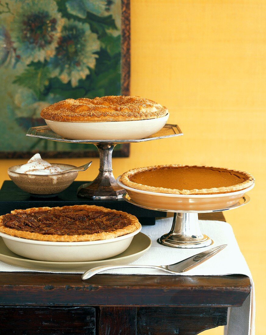 A Pecan Pie, a Squash Pie and an Apple Pie on a Wooden Table with a Bowl of Cream