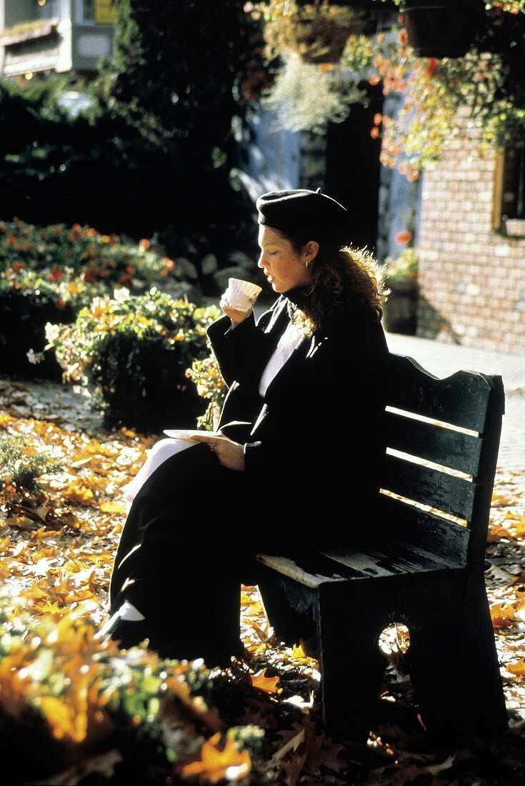 Junge Frau trinkt Tee, im Herbst auf einer Gartenbank sitzend