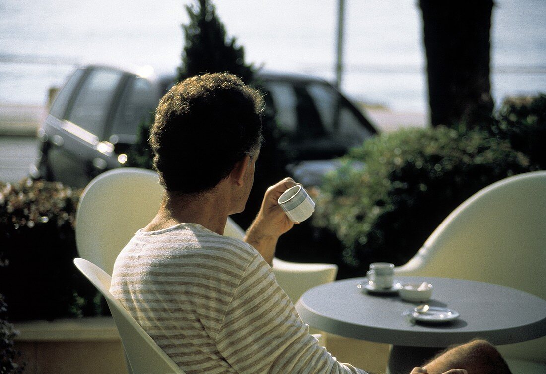 Mann sitzt am Tisch in einem Café im Freien & trinkt Kaffee