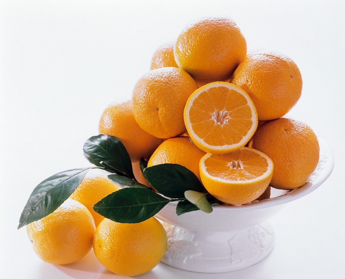 Orangen mit Zweig auf weisser Schale, eine Orange halbiert